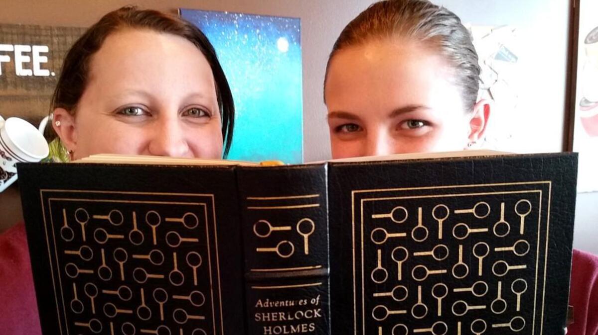 Between Friends Coffee co-owners Jaimie Miller and Victoria Hawkins smile behind a book. They announced on Tuesday, March 3, 2026, that the store will close soon.