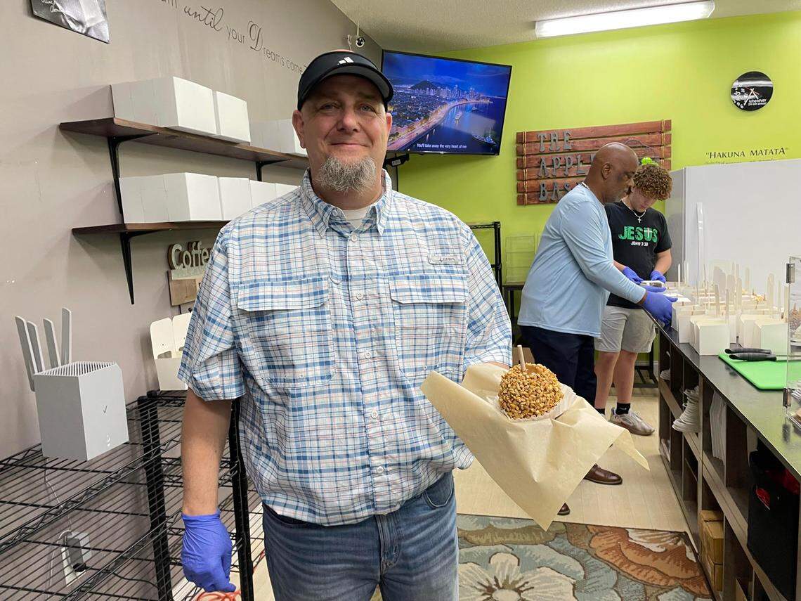Clayton Brown, who now owns The Apple Basket in Centerville with his wife, Cindy, holds a gourmet caramel apple covered with peanuts. Keith Turner, one of the original owners who taught Brown the ropes, is in the background (in blue shirt) with Brown’s son, Eli, at the reopening of the shop Monday, May 5, 2025.