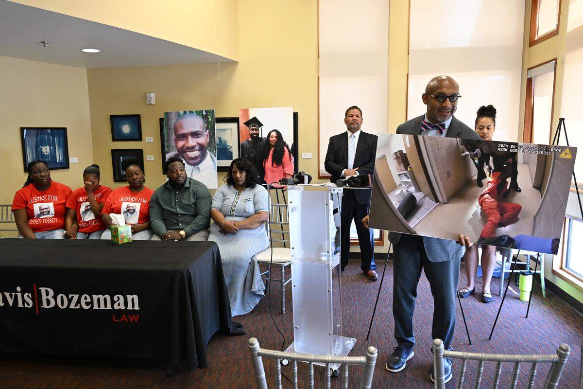 Attorney Mawuli Davis (right) holds a poster with a frame from body camera footage from the day Stephen Fossett died in custody at the Bibb County Jail, during a press conference on Thursday, May 21, 2026, at the Tubman Museum in Macon, Ga. Stephen Fossett’s death was ruled a homicide by the Georgia Bureau of Investigation after he died in the jail in May 2024.