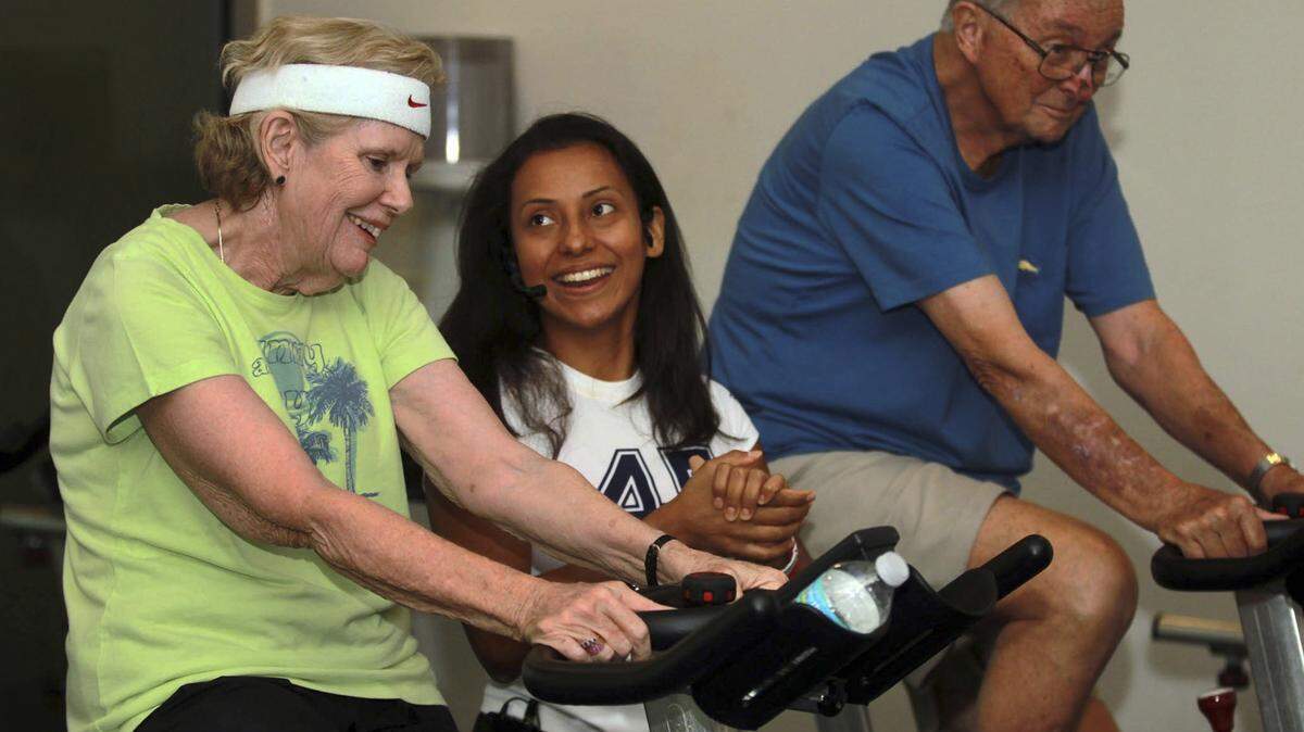 Angela Alvarado, a health coach instructor, center, helps Claire Hackett, left, and her husband, Bob, during a cycling class for individuals with Parkinson’s disease last month in Florida.