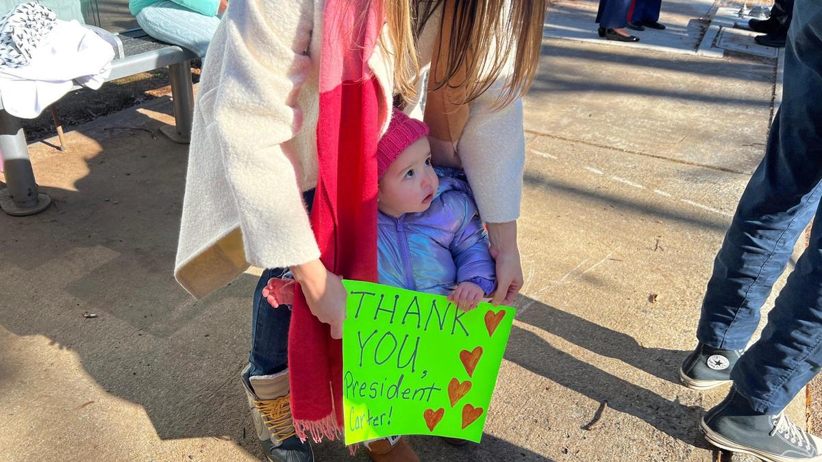 Atlanta resident Theresa Gonzalez holds up a “Thank You, President Carter!” sign for her one-year-old daughter Isabella after the hearse carrying former president Jimmy Carter departs the Carter Presidential Center on Tuesday, Jan. 7, 2024, at the Carter Presidential Center in Atlanta, Georgia. The Carter hearse and motorcade left the Carter Presidential Center and drove to Dobbins Air Reserve to fly to Washington D.C., where the former president will lie in state at the U.S. Capitol until Thursday.