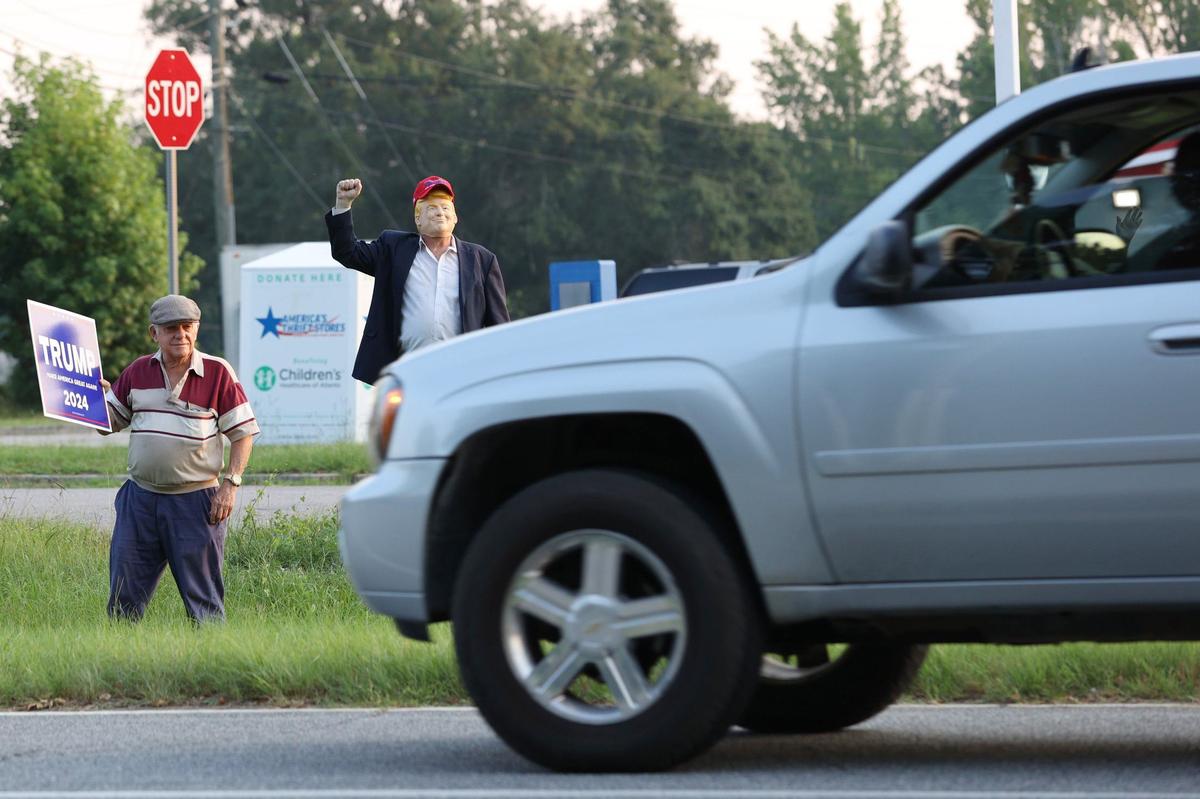 Tom Brown (left) and David Sumrall in a Donald Trump mask react to a honking car on Eisenhower Parkway on Monday, July 15, 2024, in Lizella, Georgia. A half dozen supporters, including members of the Bibb County Republican Party, gathered to show their support for former president Trump two days after an attempted assassination attempt on him in Pennsylvania.