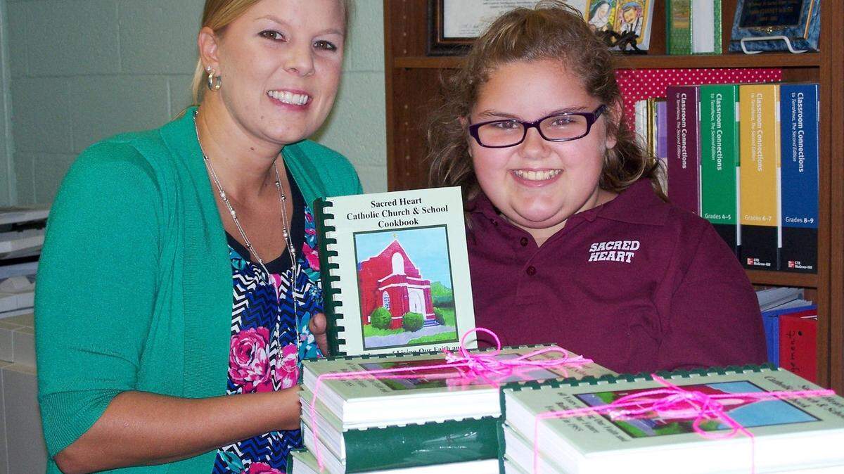 MICHAEL W. PANNELL/SPECIAL TO THE SUN NEWSAshley Lopez, president of Sacred Heart’s Home and School group, and Sacred Heart Catholic School sixth-grader Annabelle Balducci pose with the new cookbook to be sold at the fall festival. The cover features a painting by Annabelle of Sacred Heart’s former sanctuary.