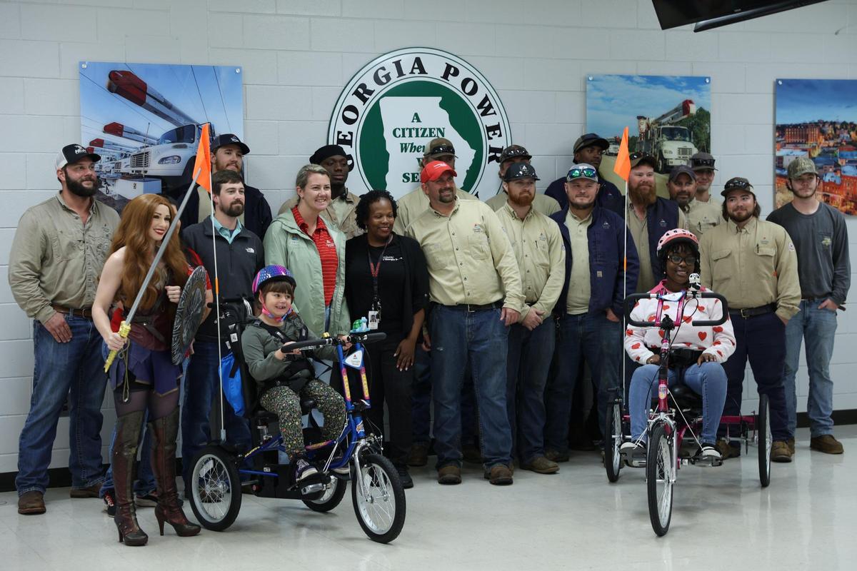 Stella Promesti-Adamek (left) and Karrington Starks pose on their new adaptive bicycles with Georgia Power employees and a woman portraying Wonder Woman on Tuesday, Nov. 19, 2024, at Georgia Power Company in Macon, Georgia. Georgia Power donated both bicycles, which were designed by Freedom Concepts.