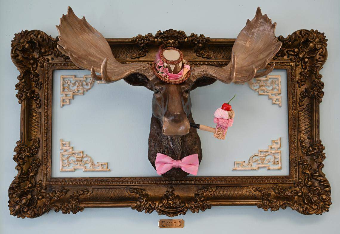A framed moose statue wearing a small top hat and smoking an ice cream pipe on the wall inside of the new Mad Hatter Cafe & Confections in Warner Robins. Various animals— most wearing hats— hang on the cafe walls along with other unique decor.