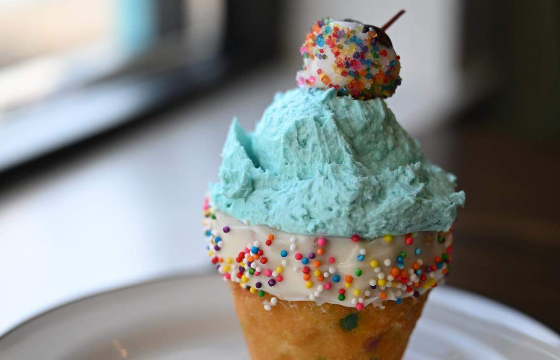 A confetti ice cream cone cake from Mad Hatter Cake & Confections in Warner Robins features pound cake shaped as an ice cream cone with confetti cake inside and covered in frosting with a chocolate covered cherry on top.