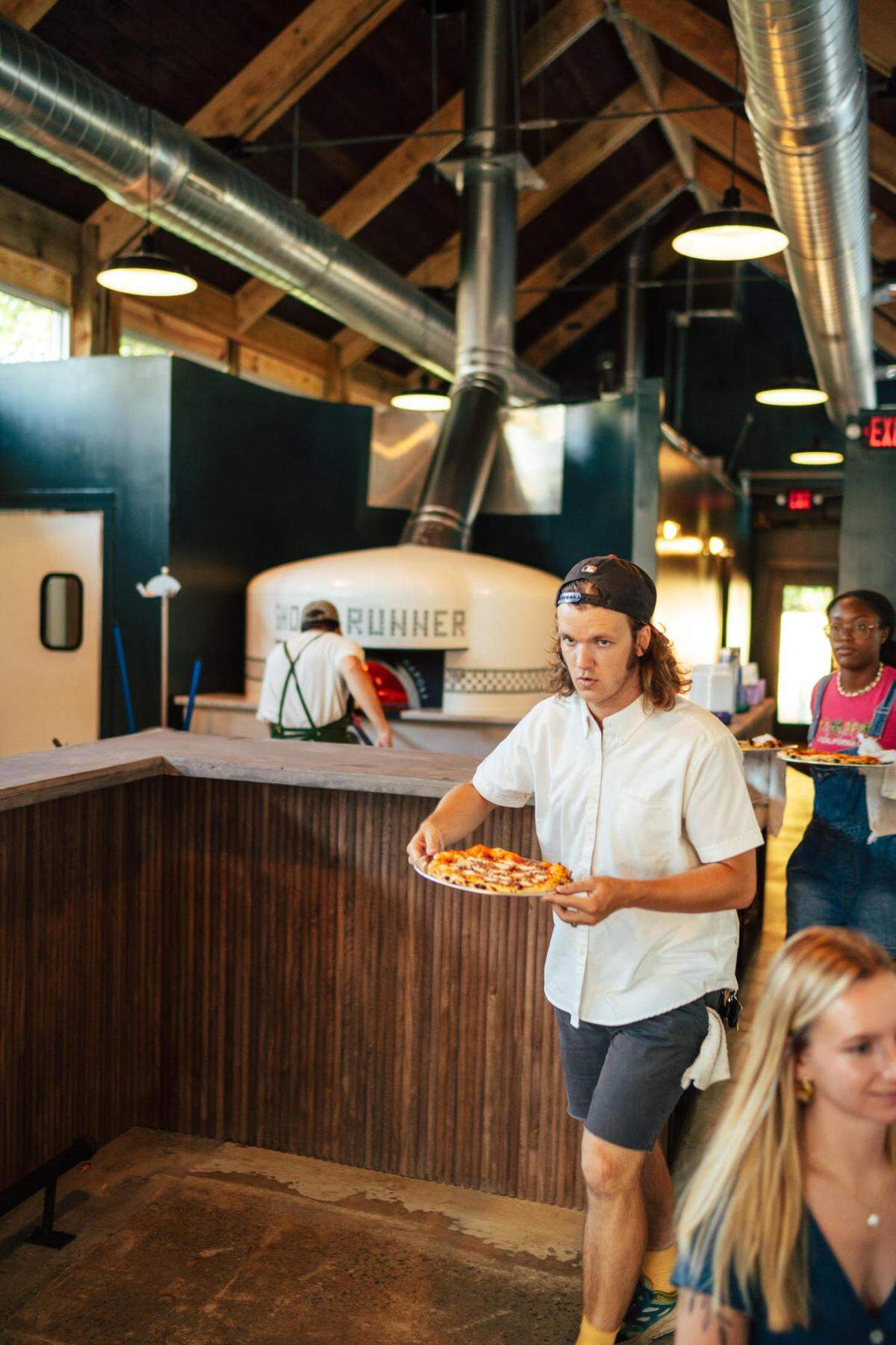 Co-owner Cody Walker takes a fresh-hot pizza to a table at the new Ghost Runner Pizza in historic downtown Perry. His brother and co-owner Wade Walker mans the Fiero Forni wood-fired brick oven in the background..