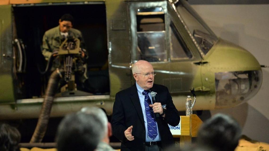 Retired Col. Robert G. Certain, with a backdrop of a Vietnam War-era helicopter gunner, tells a POW-MIA Day Recognition Day audience about his time in captivity after he and the bomber crew he served with went down over North Vietnam.