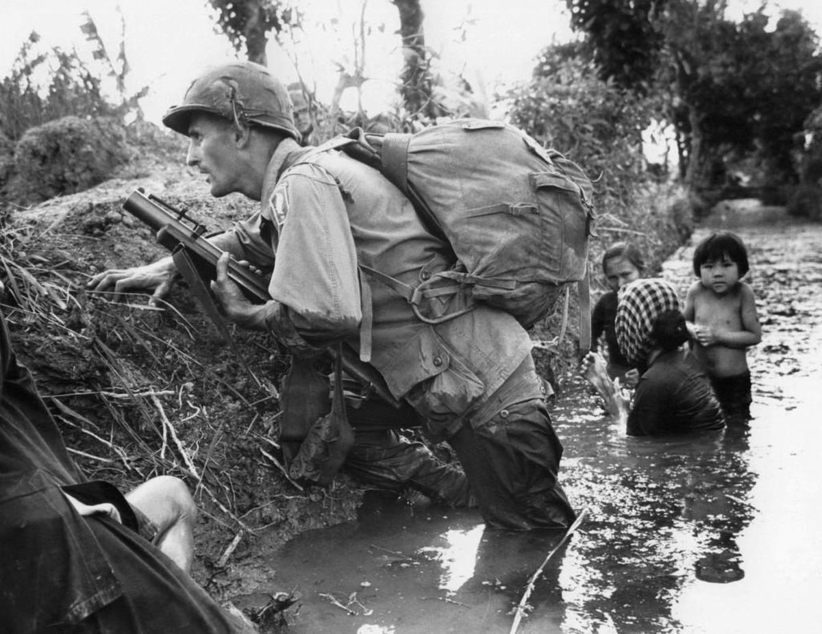 In this Jan. 1, 1966 file photo, a Paratrooper of the 173rd U.S. Airborne brigade crouches with women and children in a muddy canal as intense Viet Cong sniper fire temporarily pins down his unit during the Vietnamese War near Bao trai in Vietnam. Filmmaker Ken Burns said he hopes his 10-part documentary about the war could serve as sort of a vaccine against some problems that took root during the conflict, such as a lack of civil discourse in America.